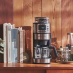 Stainless coffee maker on a wooden counter with books and a tray of mugs beside a vase of leaves against a wood paneled wall