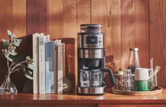 Stainless coffee maker on a wooden counter with books and a tray of mugs beside a vase of leaves against a wood paneled wall