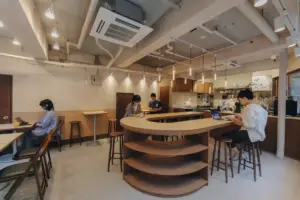 a87e392163dcca1576c2c10ae98cd5c5 | コーヒーステーション Cafe interior with a curved wooden counter and shelves several patrons sitting at tables and stools using laptops and drinks visible on the counter area behind the bar