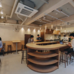 Café interior with a curved wooden bar customers at laptops warm pendant lights and an open kitchen in the background