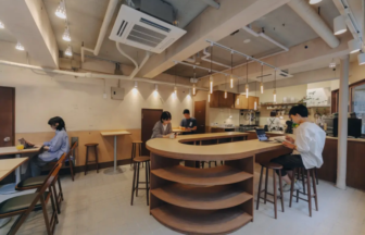 Café interior with a curved wooden bar, customers at laptops, warm pendant lights, and an open kitchen in the background.