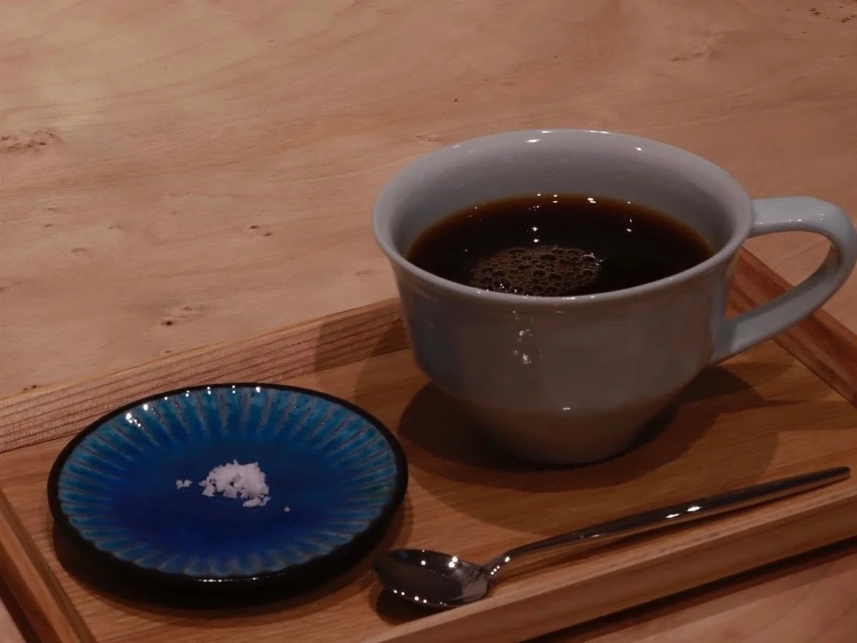 White ceramic cup of black coffee on a wooden tray with a small blue dish containing sugar and a spoon