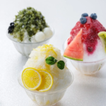 Three glass bowls of shaved ice dessert with fruit toppings lemon slices and mint in the foreground a green teablack topped bowl in the back left and a berry fruit bowl with watermelon and kiwi on the right