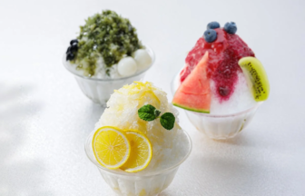 Three glass bowls of shaved ice dessert with fruit toppings lemon slices and mint in the foreground a green teablack topped bowl in the back left and a berry fruit bowl with watermelon and kiwi on the right