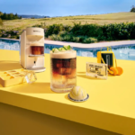 Poolside yellow counter with a white espresso machine a tall glass of iced coffee and an ice tray on a sunny day by a pool text reads Yuzu Vanilla Over Ice