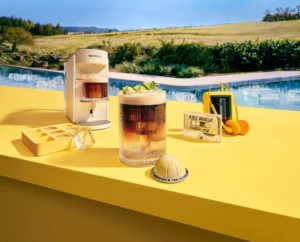Poolside yellow counter with a white espresso machine a tall glass of iced coffee and an ice tray on a sunny day by a pool text reads Yuzu Vanilla Over Ice