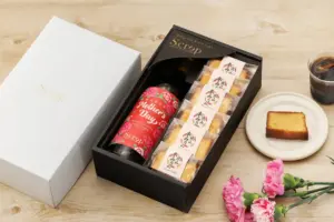 Gift set in a black box with a red branded bottle and individually wrapped snacks presented beside a white lid and a plate with cake