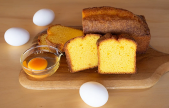 Golden yellow loaf cake sliced on a wooden cutting board with a bowl containing a raw egg and several whole eggs nearby