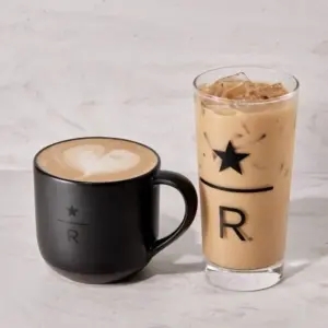 Black ceramic mug with latte art next to a tall iced coffee with a star and R logo on a light marble surface