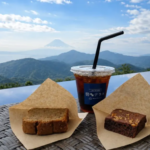 Iced coffee in a plastic cup with a black straw flanked by two pieces of cake on parchment on a wicker table with a mountain view
