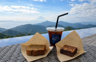 Iced coffee in a plastic cup with a black straw, flanked by two pieces of cake on parchment, on a wicker table with a mountain view.