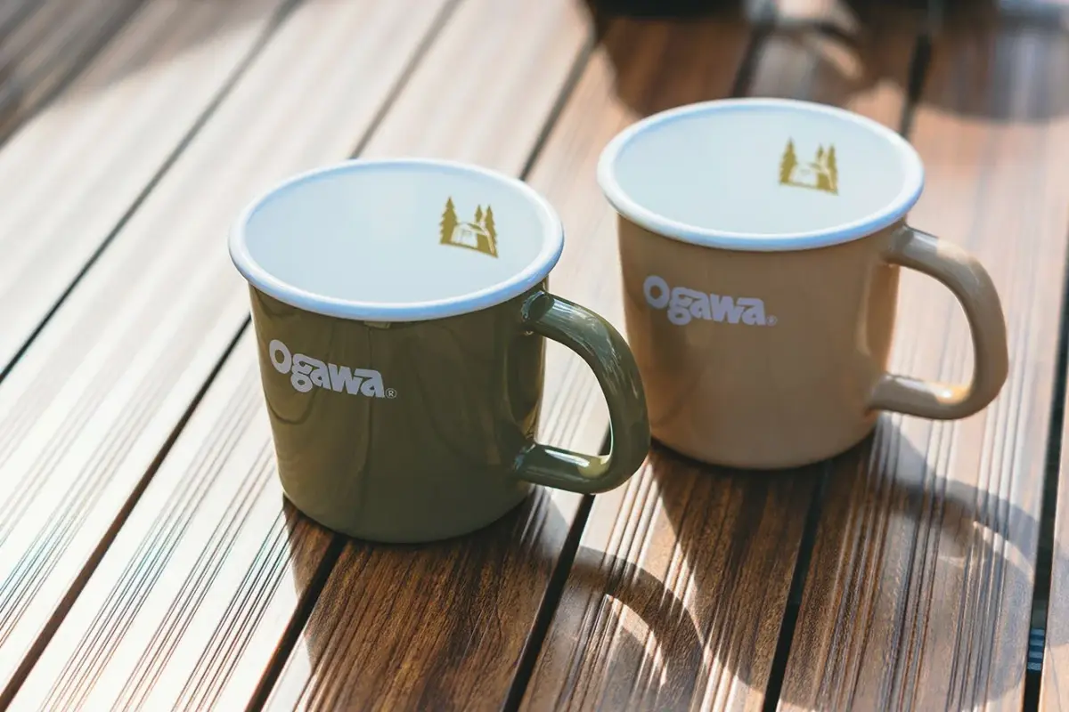 Two ceramic mugs with Ogawa logo sit on a wooden slatted table side by side in soft sunlight