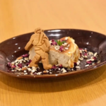 Dessert plate with a cake slice and a small cookie figure perched on top surrounded by chopped nuts and edible petals