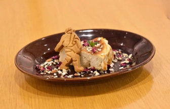 Dessert plate with a cake slice and a small cookie figure perched on top surrounded by chopped nuts and edible petals