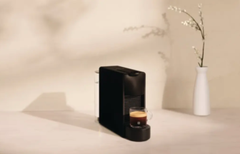 Compact black espresso machine on a light counter with a cup of coffee brewing beside it and a white vase in the background.