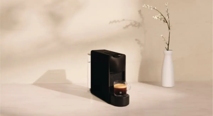 Compact black espresso machine on a light counter with a cup of coffee brewing beside it and a white vase in the background