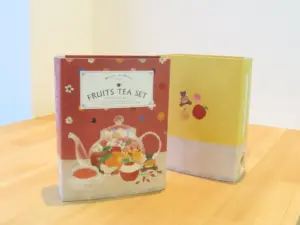 Two colorful boxes for a FRUITS TEA SET on a light wooden table one red with a teapot illustration and fruit motifs the other yellow