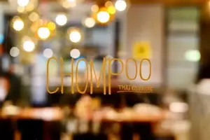Glass window of a Thai restaurant with orange lettering spelling CHOMPOO and blurred warm indoor lights in the background