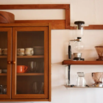 Wooden cabinet with glass doors containing dishes coffee gear on a shelf to the right