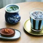 Blue and white ceramic teacup with green tea a striped iced coffee on a gold saucer and a small brown cookie on a plate on a wooden table