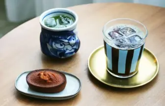 Blue-and-white ceramic teacup with green tea, a striped iced coffee on a gold saucer, and a small brown cookie on a plate on a wooden table.