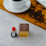 Two tiny display bottles one with a red cap and a small label the other a square brown box with yellow liquid placed on a light surface a wooden board coffee beans and a white cup are blurred in the background