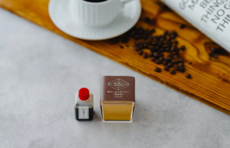 Two tiny display bottles (one with a red cap and a small label, the other a square brown box with yellow liquid) placed on a light surface; a wooden board, coffee beans, and a white cup are blurred in the background.