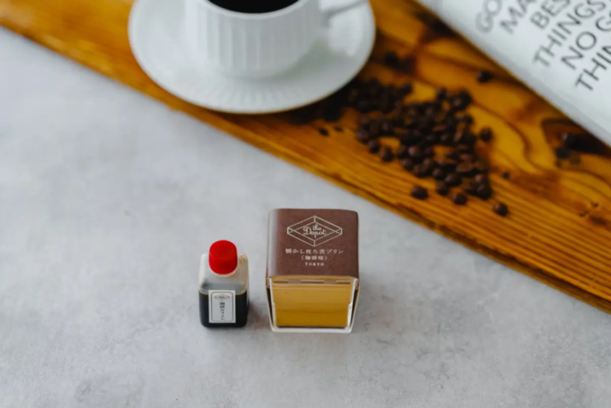 Two tiny display bottles one with a red cap and a small label the other a square brown box with yellow liquid placed on a light surface a wooden board coffee beans and a white cup are blurred in the background
