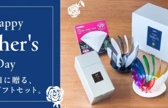 Left a dark blue card reading Happy Fathers Day with white floral corners and Japanese text right a wooden table displaying Fathers Day gifts including white gift boxes a blue Fathers Day card and a colorful stand with rainbow colored pieces arranged as a gift set