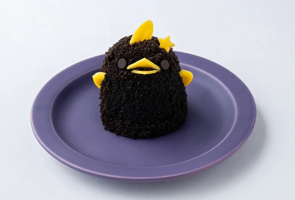 白い背景の紫色の皿に置かれた、チョコレートクランブルで覆われた鳥の形をした可愛らしいケーキです。黄色のくちばしと羽、そして頭には星形の飾りがあります。