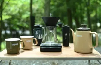 Outdoor coffee setup on a wooden table with mugs, a glass pour-over carafe on a scale, a kettle, and a grinder in a forest setting.