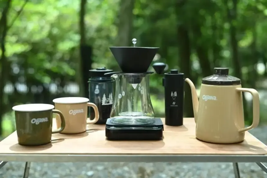 Outdoor coffee setup on a wooden table with mugs a glass pour over carafe on a scale a kettle and a grinder in a forest setting