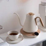White kettle with a wooden handle on a round wooden base beside a cup of tea on a clean white counter