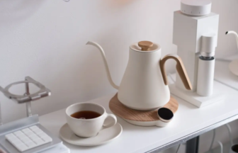 White kettle with a wooden handle on a round wooden base, beside a cup of tea on a clean white counter.