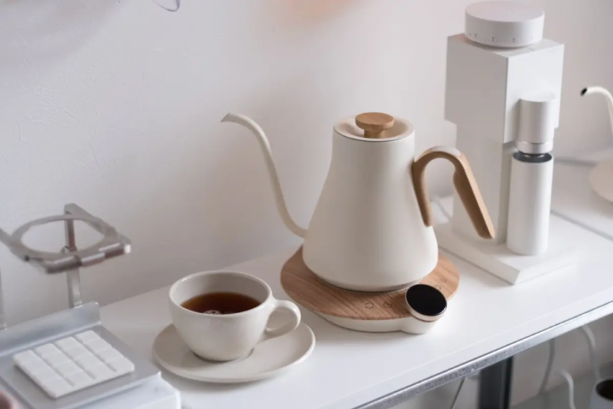 White kettle with a wooden handle on a round wooden base beside a cup of tea on a clean white counter