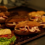Gloved hand arranging ham and cheese sliders on a baking sheet in a kitchen