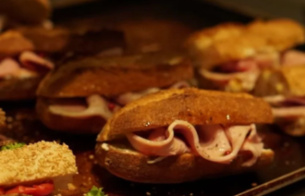 Gloved hand arranging ham and cheese sliders on a baking sheet in a kitchen.