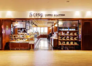 eda93dab4cb521fd6311cebd7ab572d1 | コーヒーステーション Scrop Coffee Roasters cafe interior featuring a pastry display warm wood fixtures and shelves of bags and mugs along the right wall