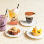 Assorted café treats on a light table two colorful smoothies a black coffee with cocoa a slice of loaf cake and a mango tart with ice cream on separate plates