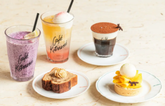 Assorted café treats on a light table: two colorful smoothies, a black coffee with cocoa, a slice of loaf cake, and a mango tart with ice cream on separate plates.