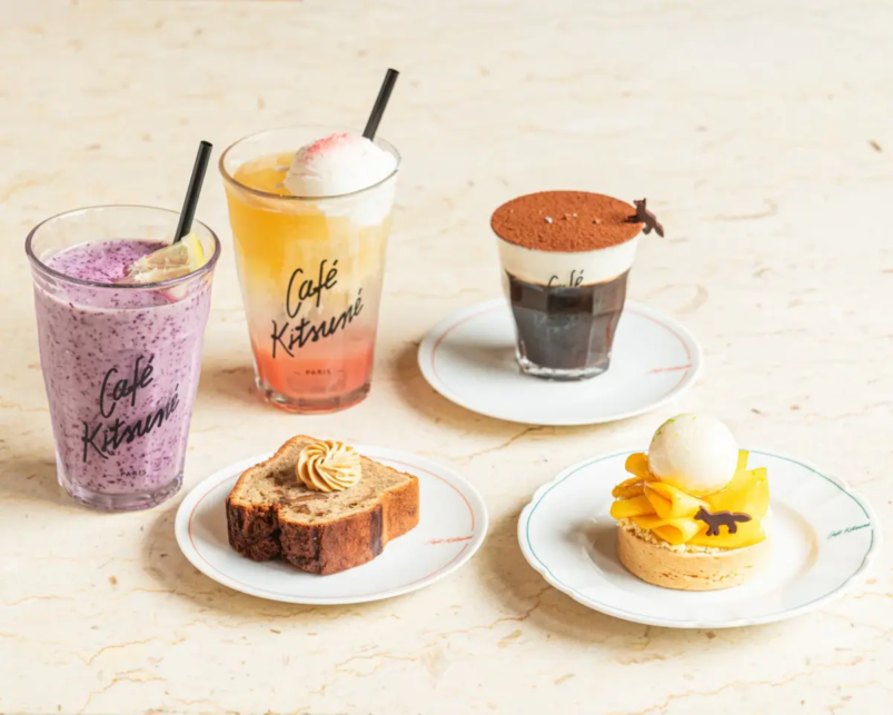 Assorted café treats on a light table two colorful smoothies a black coffee with cocoa a slice of loaf cake and a mango tart with ice cream on separate plates