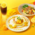 Bright yellow background with a dessert plate pancake with mango and caramel on a MARFA plate mango slices a lemon a fresh herb garnish and two fruit drinks beside a veggie pasta dish and peppers in the background