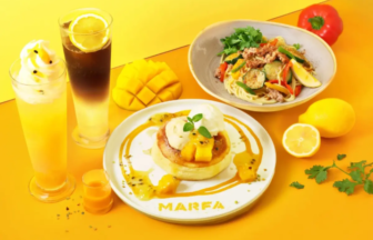 Bright yellow background with a dessert plate pancake with mango and caramel on a MARFA plate mango slices a lemon a fresh herb garnish and two fruit drinks beside a veggie pasta dish and peppers in the background