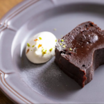 Slice of dark chocolate cake with a scoop of vanilla ice cream on a gray plate sprinkled with pistachio bits on top of the ice cream