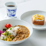 White bowl of curry with rice and assorted vegetables peas cherry tomatoes greens beside a cup of coffee and a slice of cake on a separate plate on a light table
