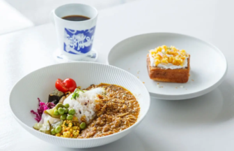 White bowl of curry with rice and assorted vegetables peas cherry tomatoes greens beside a cup of coffee and a slice of cake on a separate plate on a light table
