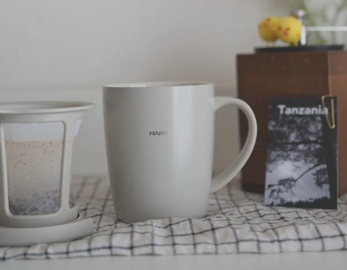 17804007972ZmR03uI9Q7dH6CsaSxKVcoi | コーヒーステーション White mug centered on a checkered table with a tea infuser to the left and a Tanzania brochure on the right
