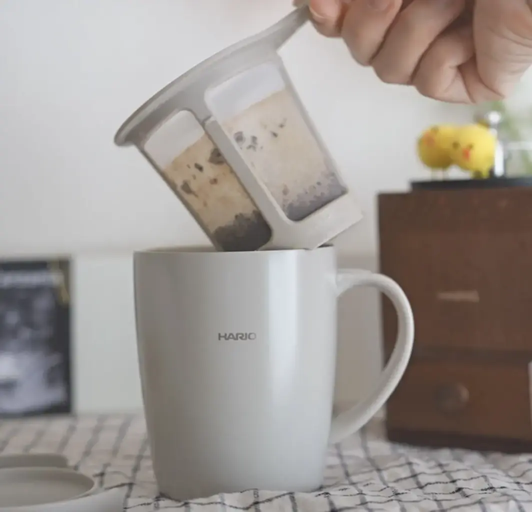 1780402348lIfDH04YTjw5qVCcRAFyk92s | コーヒーステーション Hands pour loose tea leaves from a dual chamber infuser into a white HARIO mug The scene sits on a checkered tablecloth with a wooden cabinet in the background