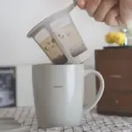 Hands pour loose tea leaves from a dual chamber infuser into a white HARIO mug The scene sits on a checkered tablecloth with a wooden cabinet in the background
