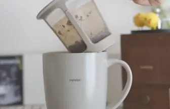 Hands pour loose tea leaves from a dual chamber infuser into a white HARIO mug The scene sits on a checkered tablecloth with a wooden cabinet in the background